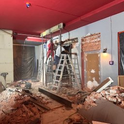 GENERAL-BUD SPÓŁKA Z OGRANICZONĄ ODPOWIEDZIALNOŚCIĄ - Dwóch pracowników na drabinach podczas wyburzania ściany z cegły w pomieszczeniu z czerwonym sufitem, gruz na podłodze. Widoczne metalowe belki wzmacniające.
