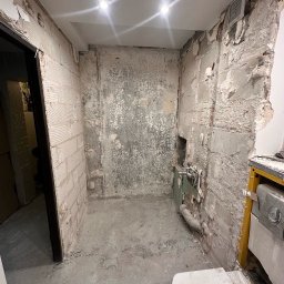 GENERAL-BUD SPÓŁKA Z OGRANICZONĄ ODPOWIEDZIALNOŚCIĄ - Zdjęcie wnętrza łazienki w trakcie demontażu: odsłonięte cegły, resztki płytek, instalacja wodna. Widoczne elementy armatury i fragment toalety.