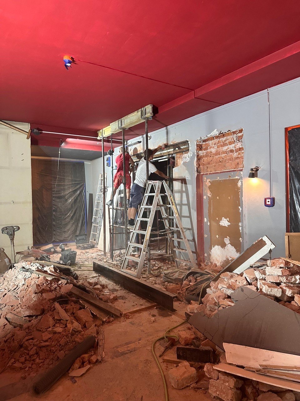 Dwóch pracowników na drabinach podczas wyburzania ściany z cegły w pomieszczeniu z czerwonym sufitem, gruz na podłodze. Widoczne metalowe belki wzmacniające.