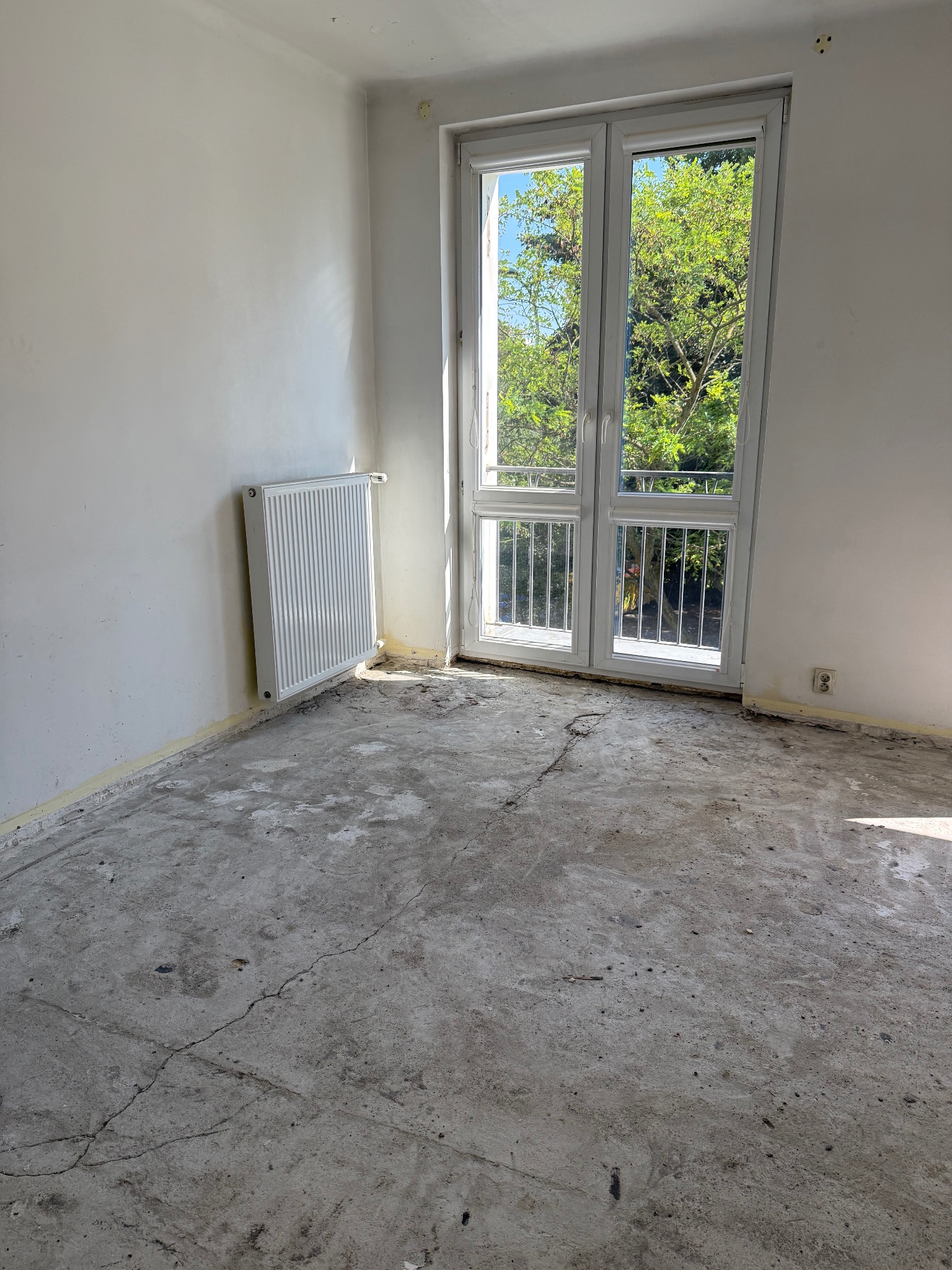 Wnętrze pokoju przed remontem: gołe ściany, grzejnik, okno balkonowe z widokiem na zieleń, surowa, popękana podłoga. Przygotowanie do prac wykończeniowych.