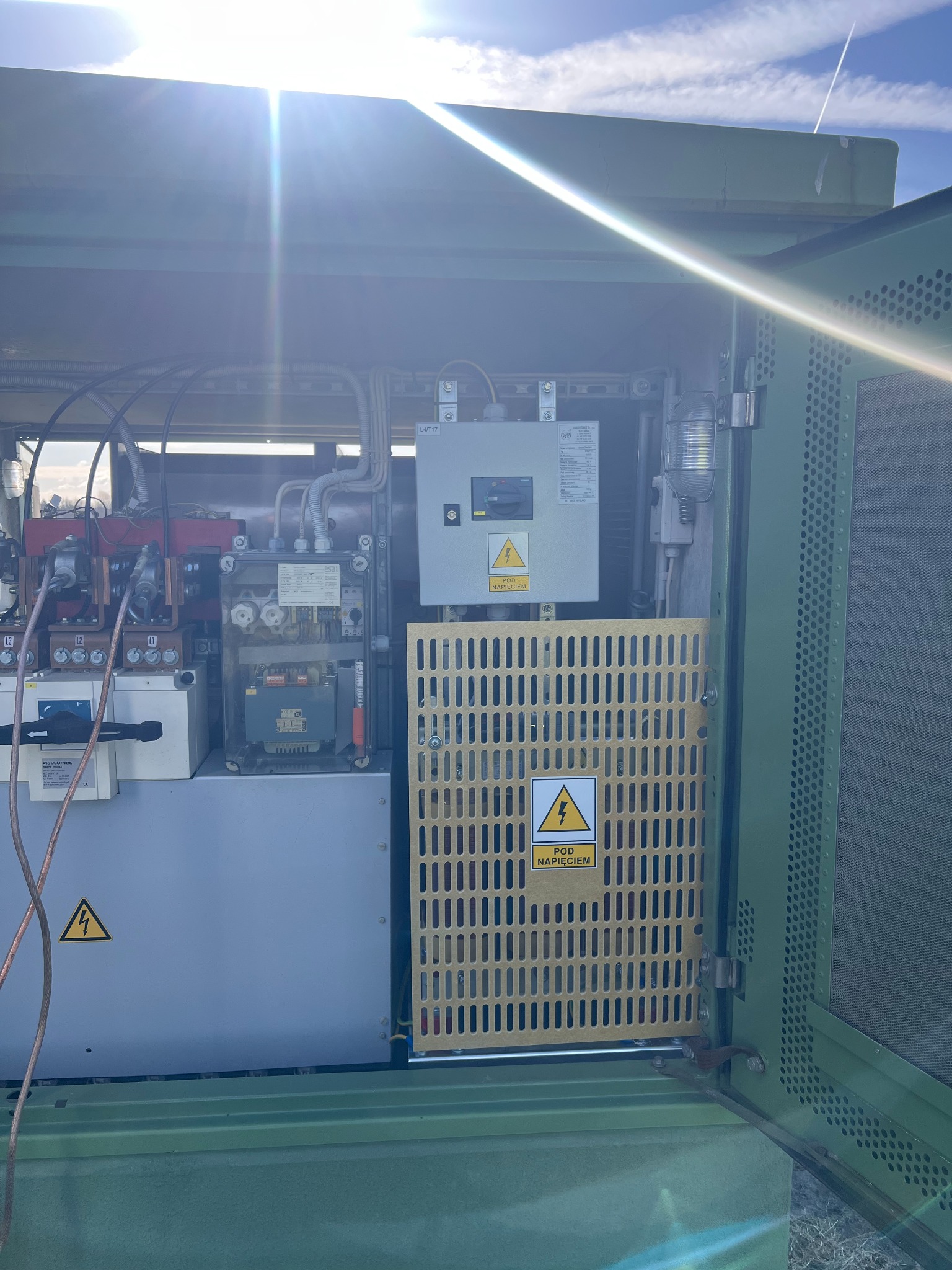 Otwarta szafa elektryczna z widocznymi elementami instalacji wewnątrz, zabezpieczona żółtą kratką z ostrzeżeniem o wysokim napięciu. Widok na farmie wiatrowej.