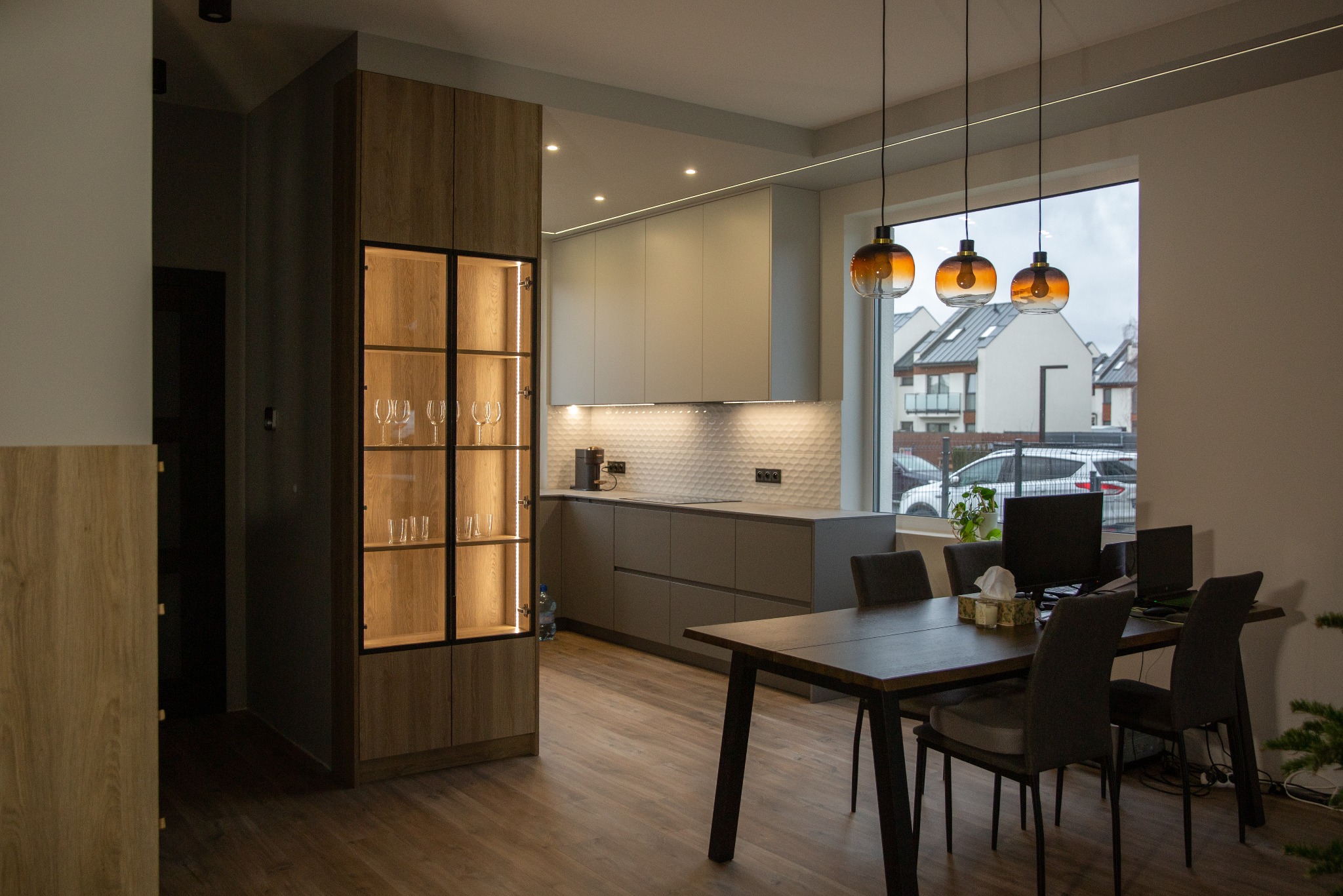 Nowoczesna kuchnia z drewnianą podłogą, szarymi szafkami i przeszkloną witryną z oświetleniem LED. Nad stołem wiszą trzy bursztynowe lampy.