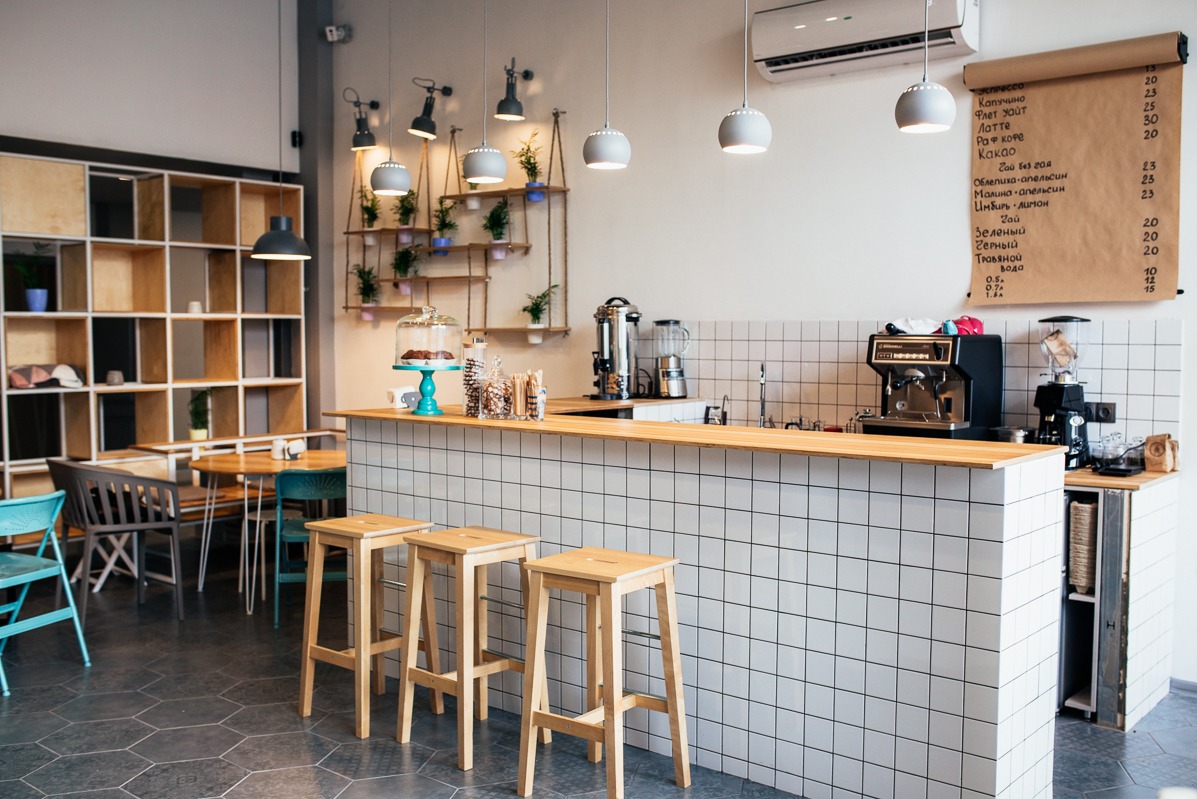 Białe kafelki kwadratowe zdobią bar w kawiarni. Drewniany blat, krzesła barowe, menu na ścianie i ekspres do kawy tworzą przytulny klimat.