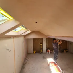 Praca przy suficie podwieszanym z płyt karton-gips w jasnym wnętrzu z oknami dachowymi. Widoczny pracownik, narzędzia i materiały budowlane.