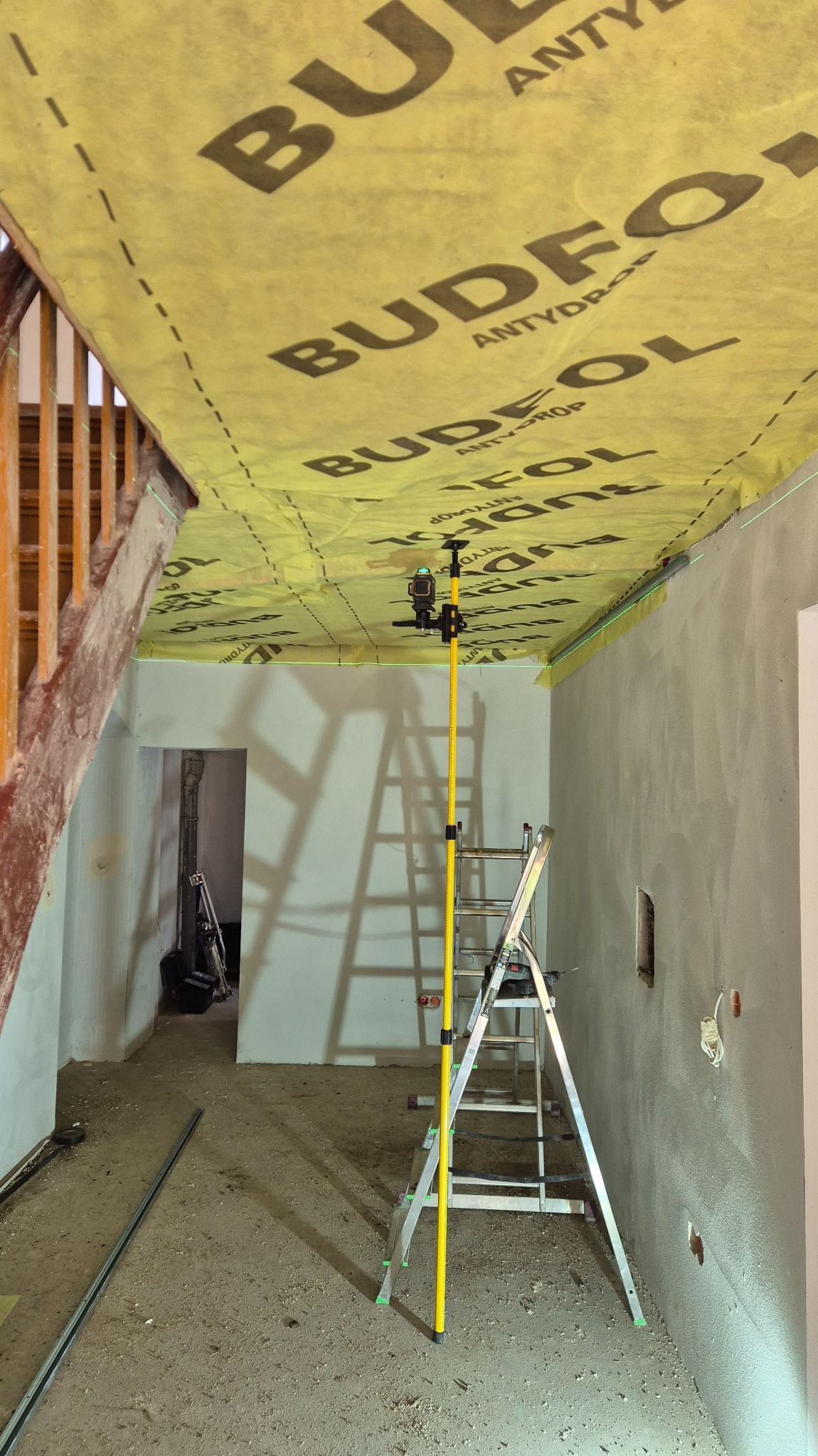 Montaż folii paroizolacyjnej Budfol na suficie podwieszanym. Widoczna drabina, poziomica laserowa i fragment schodów. Przygotowanie do dalszych prac wykończeniowych.