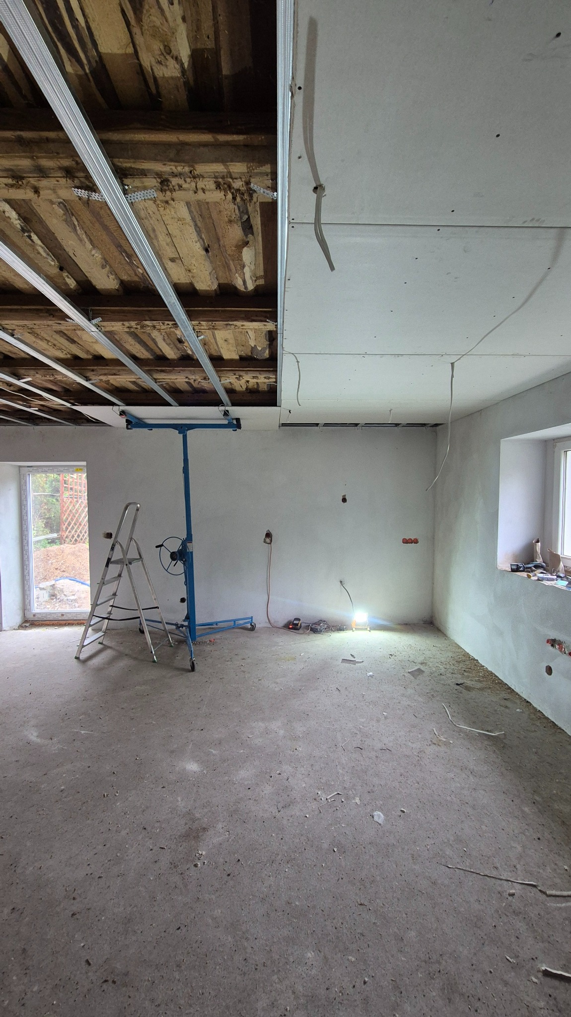 Wnętrze w trakcie montażu sufitu z płyt gipsowo-kartonowych. Widoczne profile, płyty oraz podnośnik do płyt. Okno z parapetem i schody w tle.