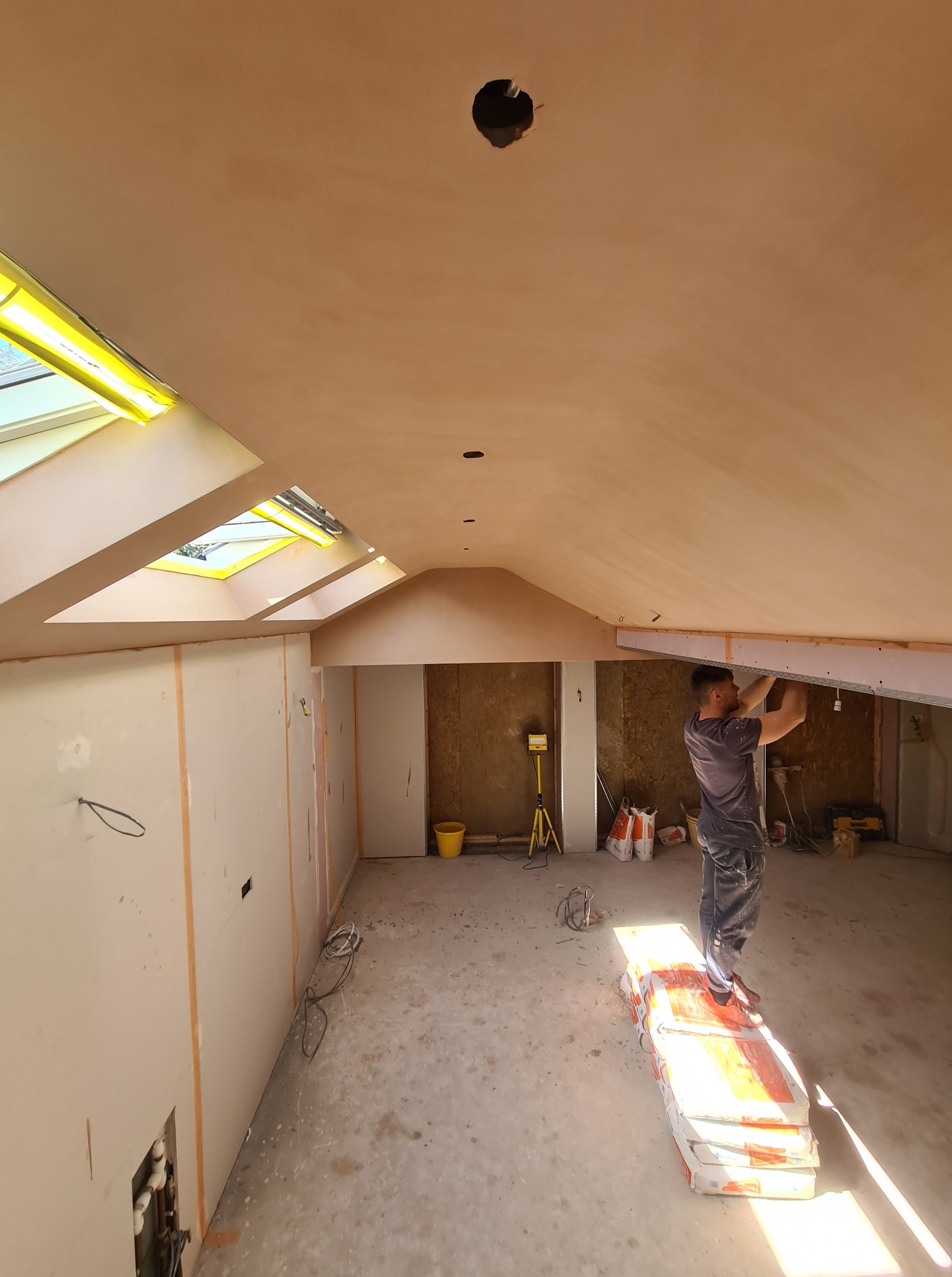 Praca przy suficie podwieszanym z płyt karton-gips w jasnym wnętrzu z oknami dachowymi. Widoczny pracownik, narzędzia i materiały budowlane.