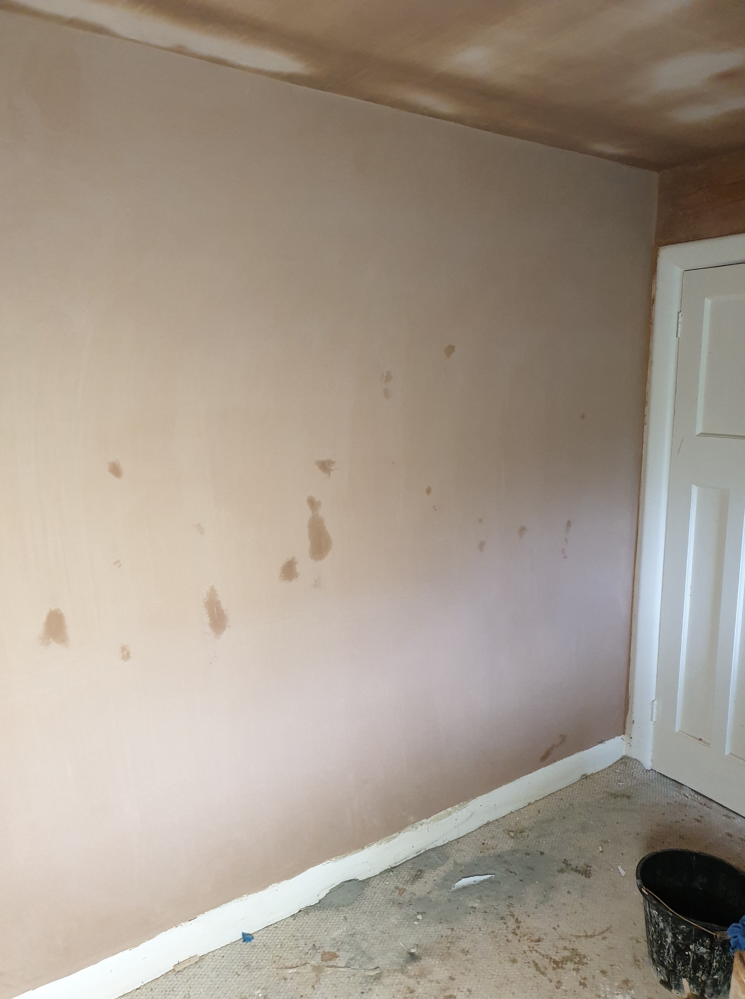 Ściana po szpachlowaniu z widocznymi plamami, przygotowana do dalszych prac wykończeniowych. W rogu widoczna biała futryna drzwi i wiadro na podłodze.