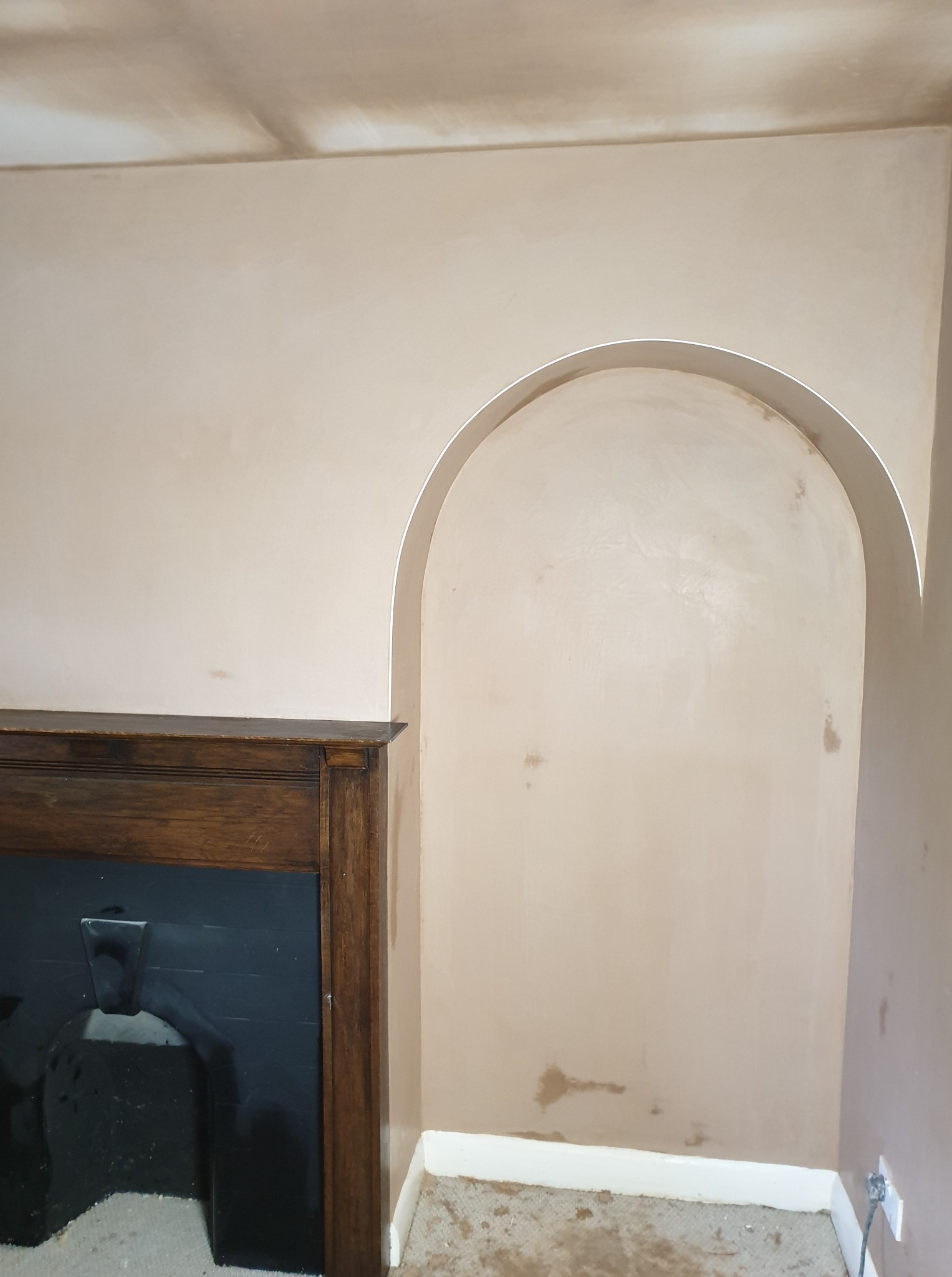 Ściana po szpachlowaniu z wnęką w kształcie łuku i fragmentem kominka. Widoczne ślady szpachli na ścianie. Prace wykończeniowe w trakcie realizacji.