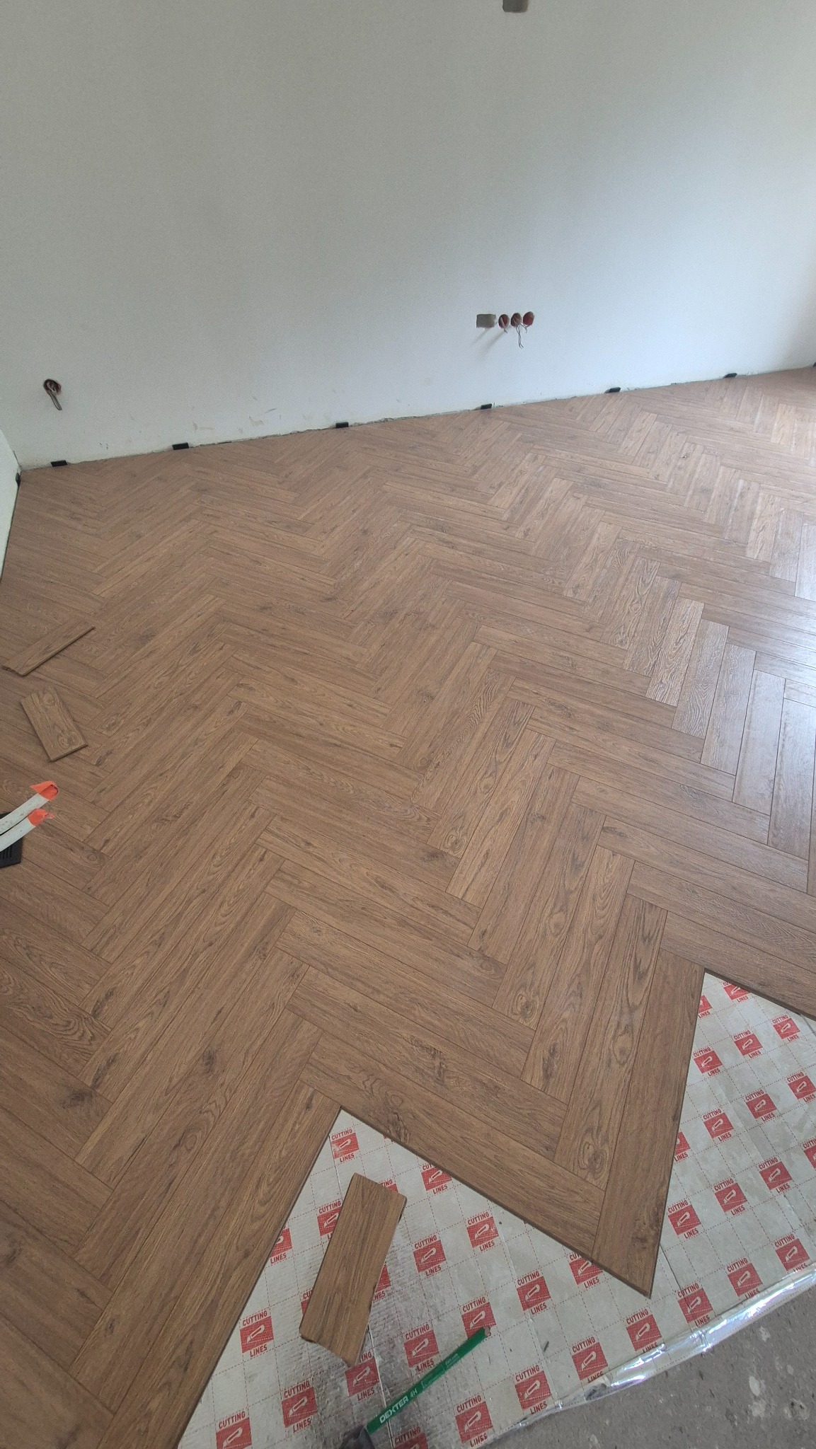 Układanie paneli podłogowych w jodełkę, widoczne narzędzia i podkład, częściowo ułożona podłoga w jasnym wnętrzu. Detal układania paneli.