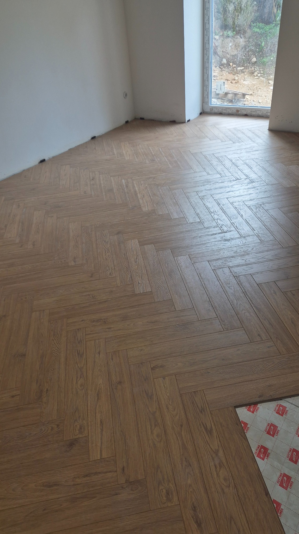 Ukończona podłoga z paneli ułożonych w jodełkę w jasnym pomieszczeniu z oknem. Fragment podkładu pod panele widoczny w rogu.