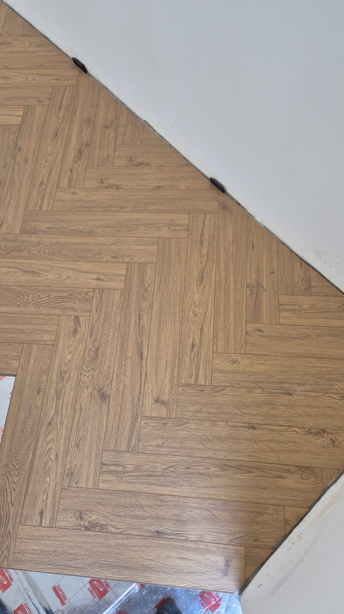 Ukończona podłoga z paneli w jodełkę, brązowe panele imitujące drewno, przy białej ścianie. Częściowo widoczne materiały montażowe w lewym dolnym rogu.