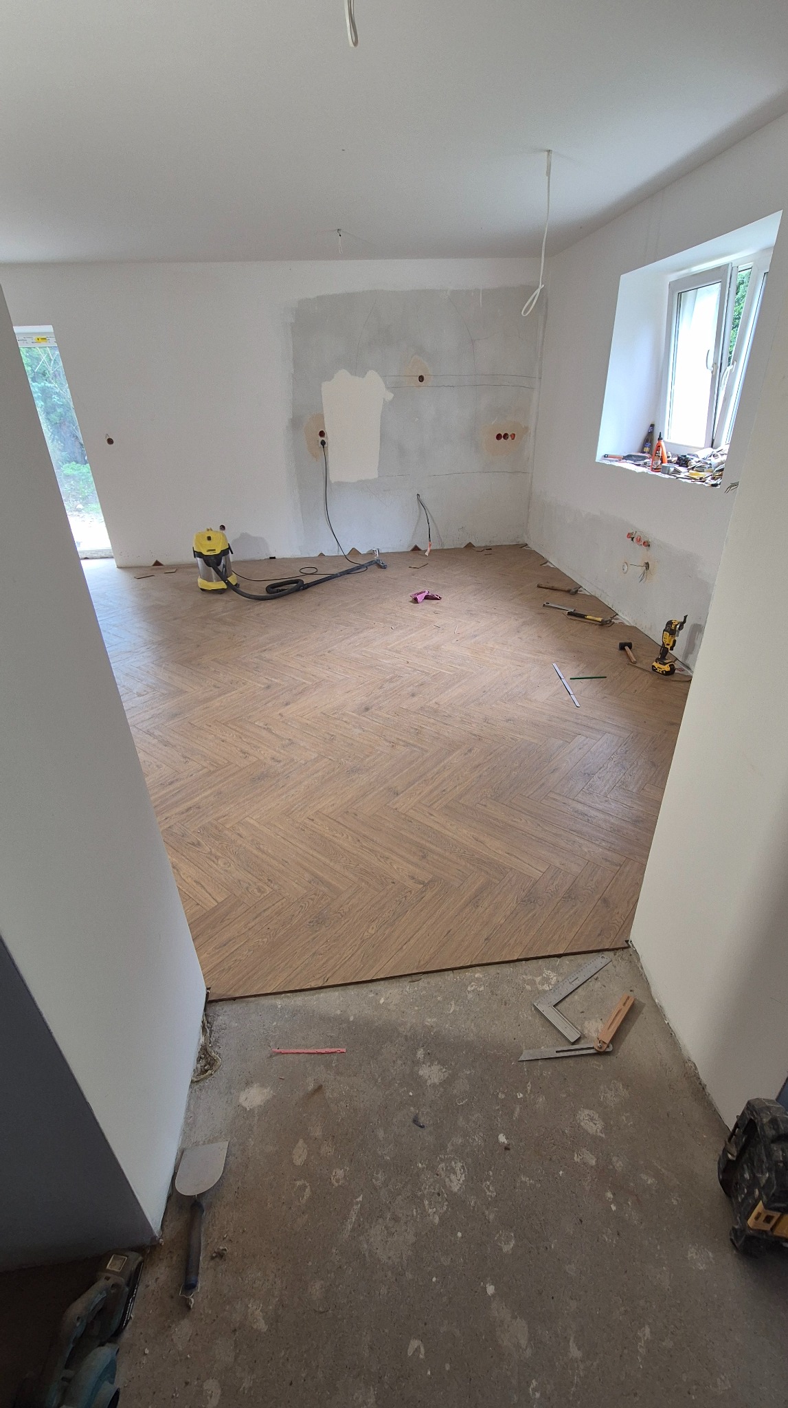 Układanie parkietu w jodełkę w jasnym wnętrzu. Widoczne narzędzia, odkurzacz i fragmenty surowych ścian. Częściowo wykończona podłoga w trakcie prac.