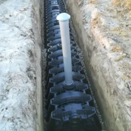 Modułowa instalacja rozsączająca w wykopie, obsypana żwirem. System drenażowy z rurą pionową i odpowietrznikiem. Widok z góry na czarne elementy w ziemi.