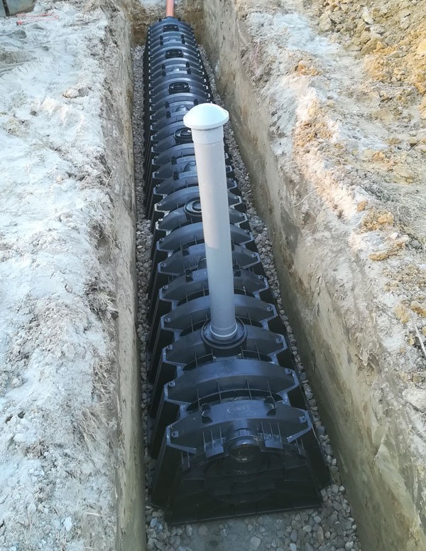 Modułowa instalacja rozsączająca w wykopie, obsypana żwirem. System drenażowy z rurą pionową i odpowietrznikiem. Widok z góry na czarne elementy w ziemi.