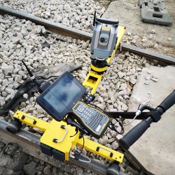 ProGeo Patryk Sadecki - Żółty wózek geodezyjny z teodolitem i tabletem na szynach kolejowych, otoczony żwirem. Widoczny fragment toru i sygnalizator.