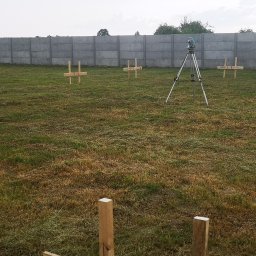 ProGeo Patryk Sadecki - Tyczenie działki: niwelator na statywie i drewniane krzyże geodezyjne na trawiastym terenie, w tle betonowy płot i drzewa. Prace pomiarowe w toku.