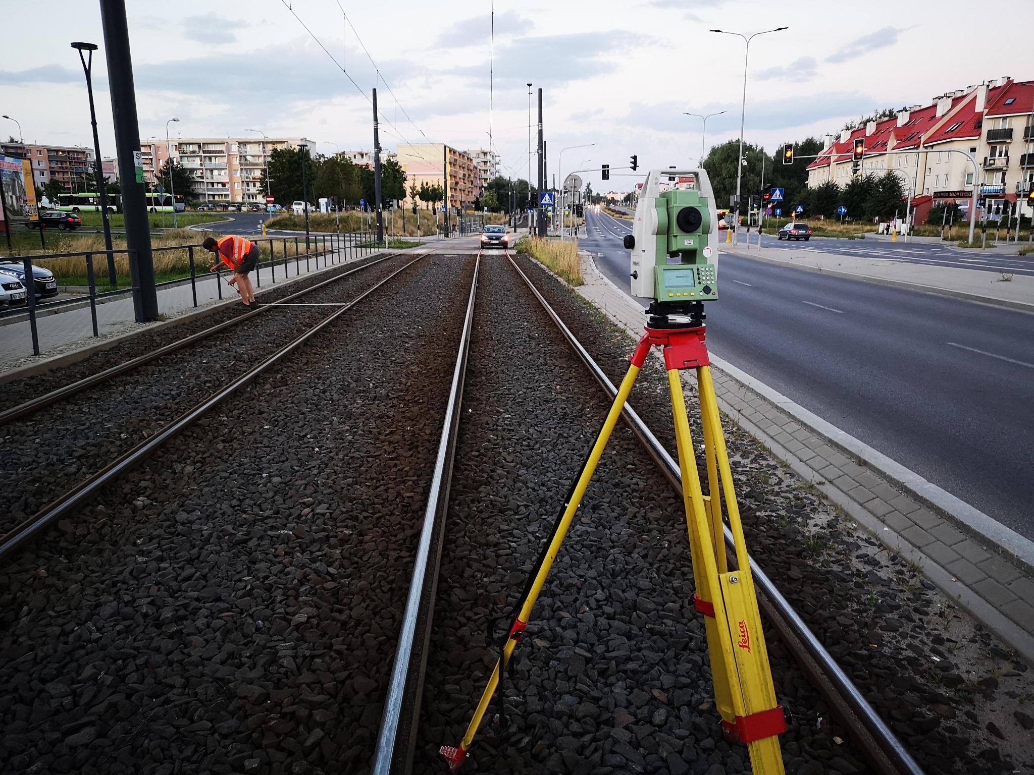 Praca pomiarowa na torach tramwajowych: żółty statyw z teodolitem Leica na tle ulicy i budynków. Osoba w pomarańczowej kamizelce przy torach, pomiary.