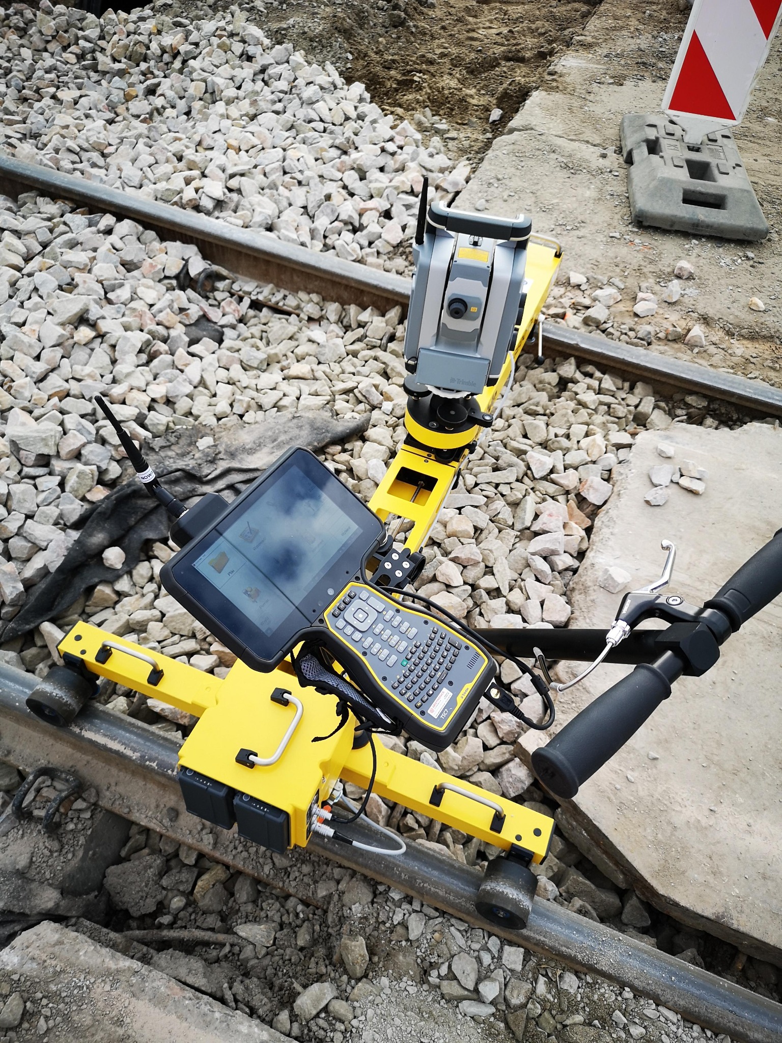 Żółty wózek geodezyjny z teodolitem i tabletem na szynach kolejowych, otoczony żwirem. Widoczny fragment toru i sygnalizator.