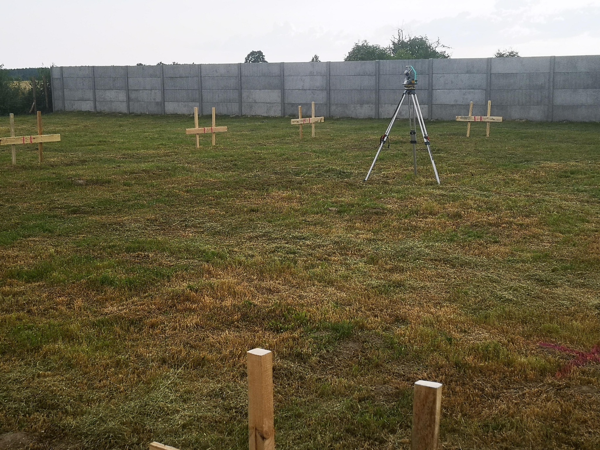 Tyczenie działki: niwelator na statywie i drewniane krzyże geodezyjne na trawiastym terenie, w tle betonowy płot i drzewa. Prace pomiarowe w toku.