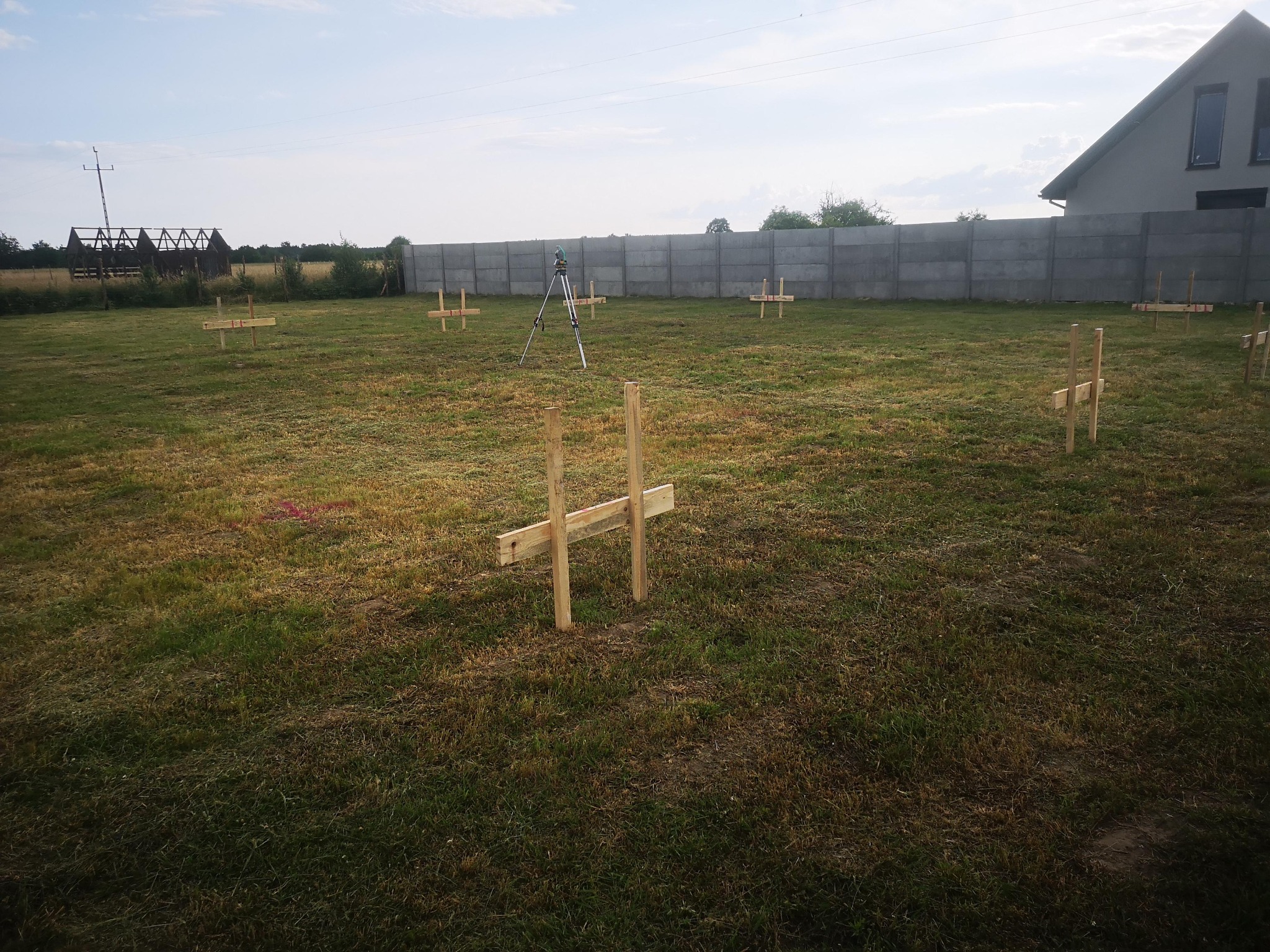 Teren po niwelacji z palikami, tyczkami i niwelatorem laserowym na trójnogu, przygotowany pod budowę domu. W tle szary betonowy płot i konstrukcja dachu.