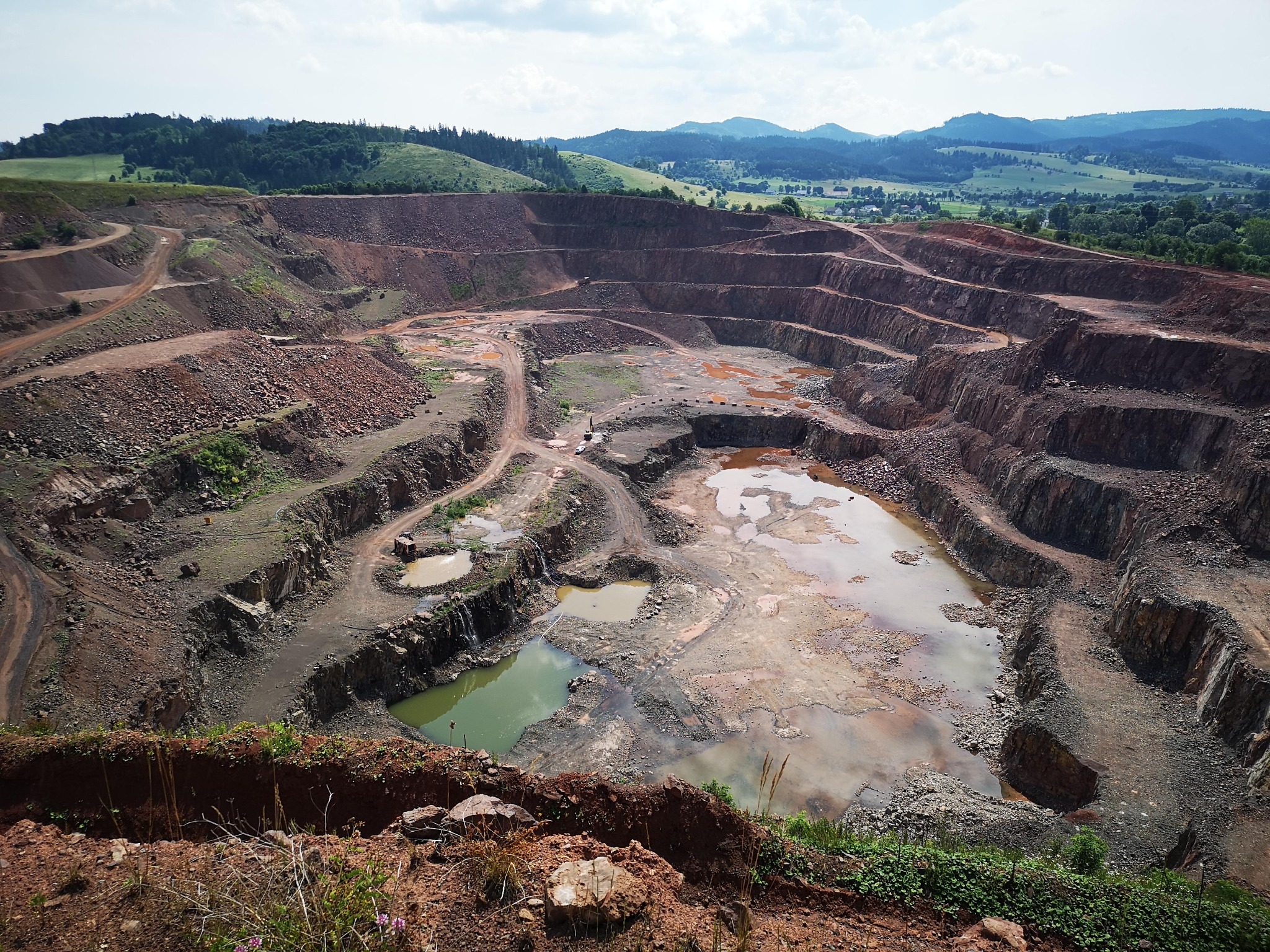 Rozległy kamieniołom z tarasowymi poziomami i zbiornikami wodnymi, w tle zalesione wzgórza pod błękitnym niebem z obłokami. Widok z góry na wyrobisko.