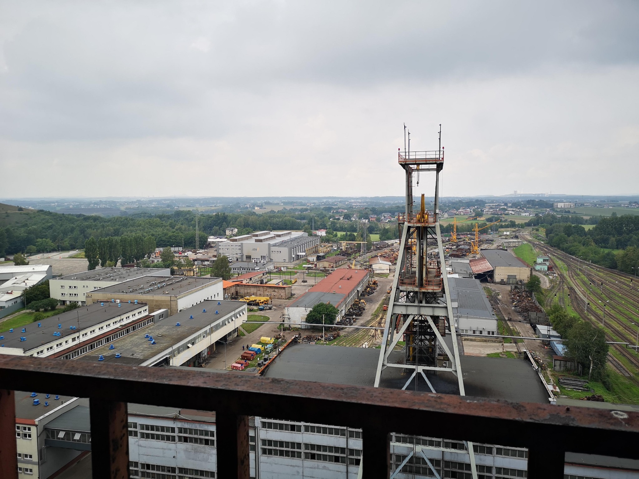 Widok z góry na teren przemysłowy z wieżą wyciągową na pierwszym planie i budynkami w tle, pod pochmurnym niebem.
