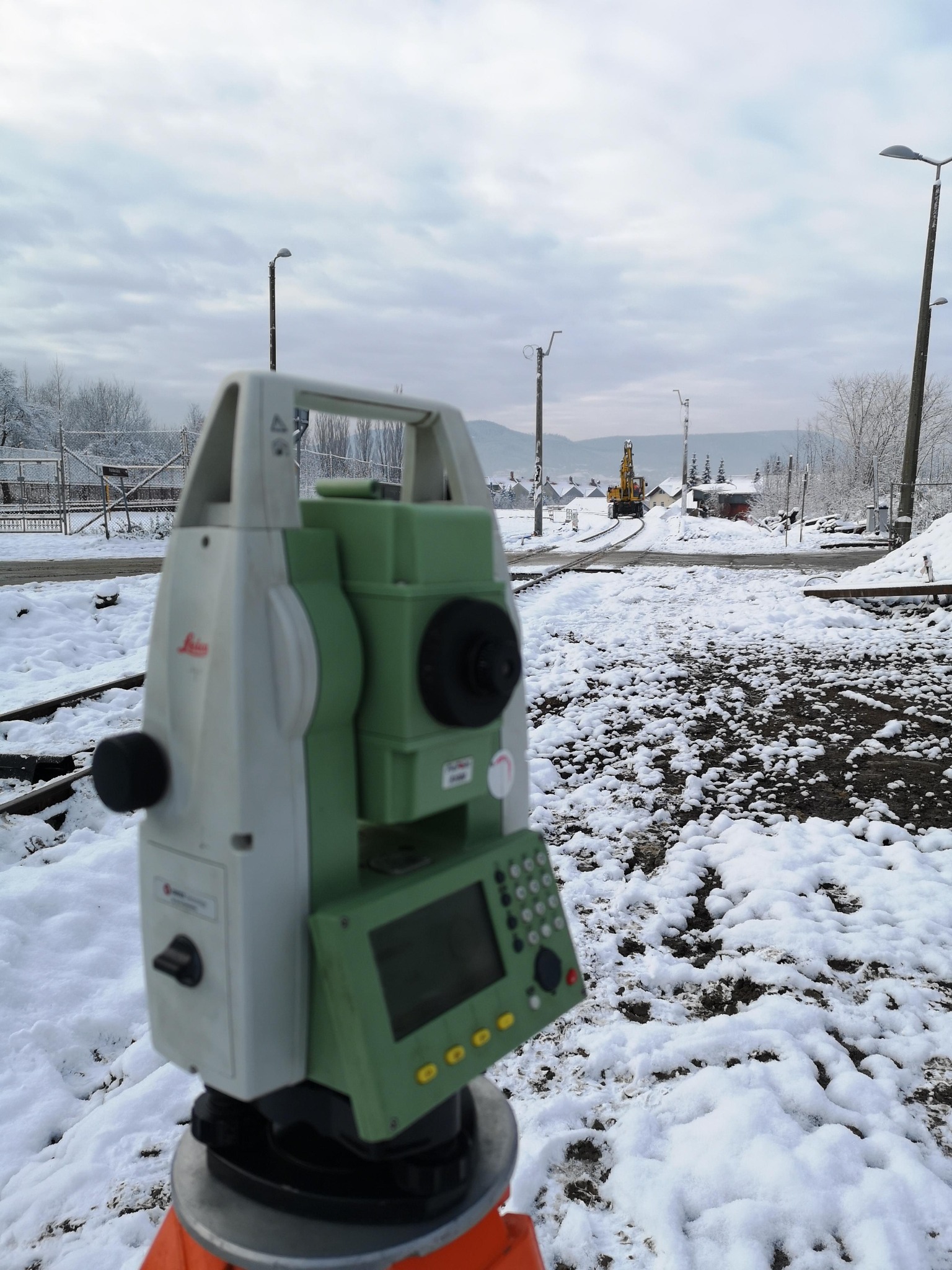 Tachimetr Leica na statywie w zimowym plenerze. W tle tor kolejowy, budowa i koparka. Pochmurne niebo.