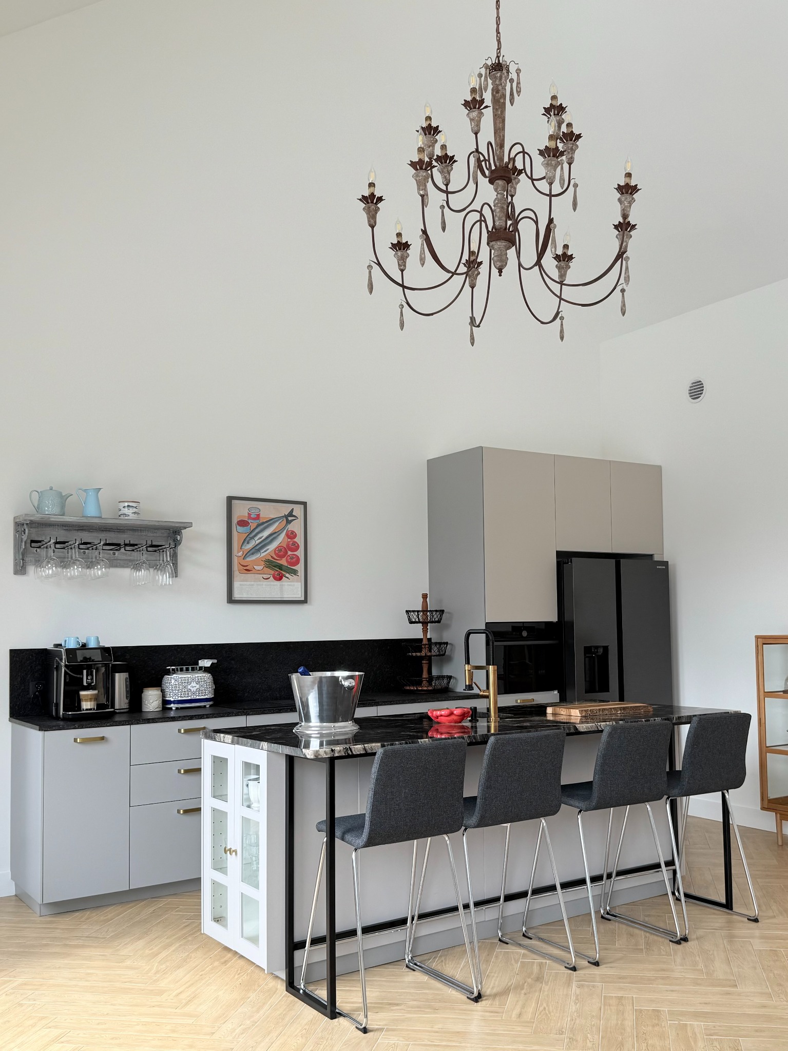 Elegancka kuchnia z wyspą, czarnym blatem i wiszącym żyrandolem. Jasne, przestronne wnętrze z drewnianą podłogą w jodełkę. Nowoczesne krzesła barowe.