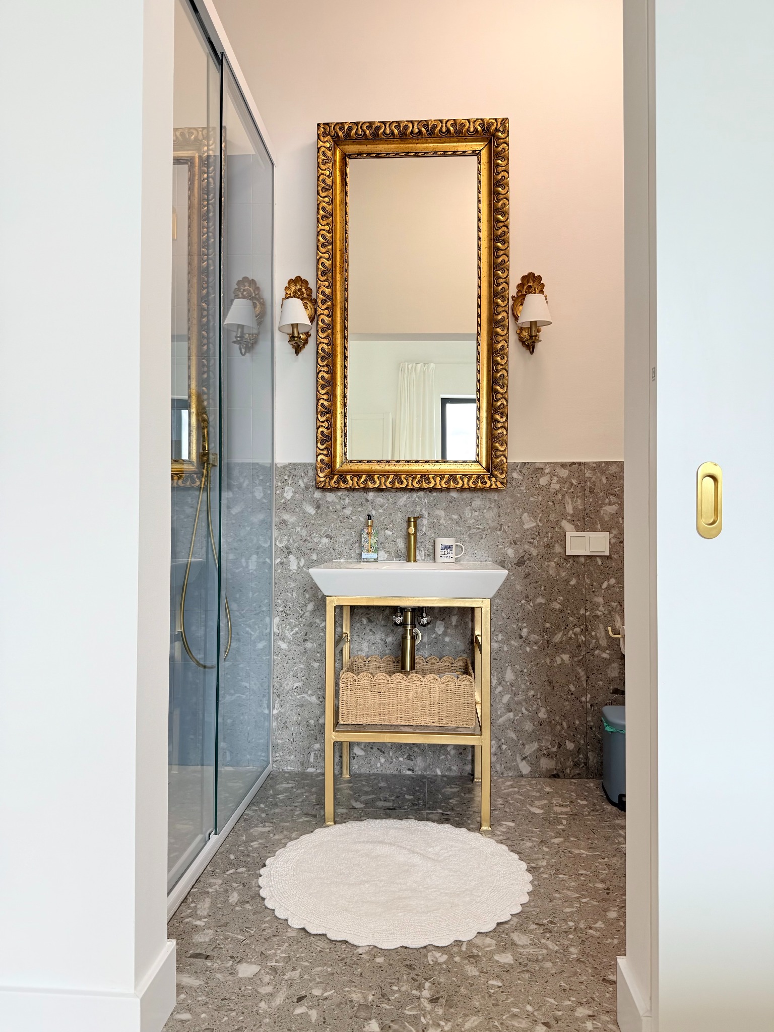 Elegancka łazienka z umywalką na złotym stelażu, ozdobnym lustrem w złotej ramie i kabiną prysznicową. Wnętrze wykończone płytkami lastryko.