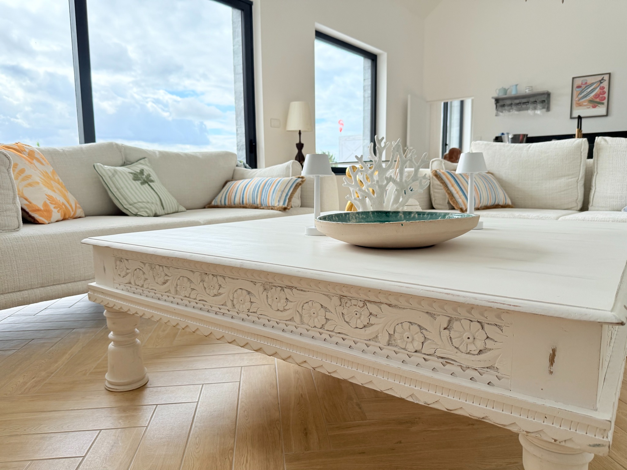 Jasne wnętrze salonu z białym narożnikiem, zdobionym stolikiem kawowym i parkietem ułożonym w jodełkę. Widoczne okna z widokiem na niebo i dekoracje.