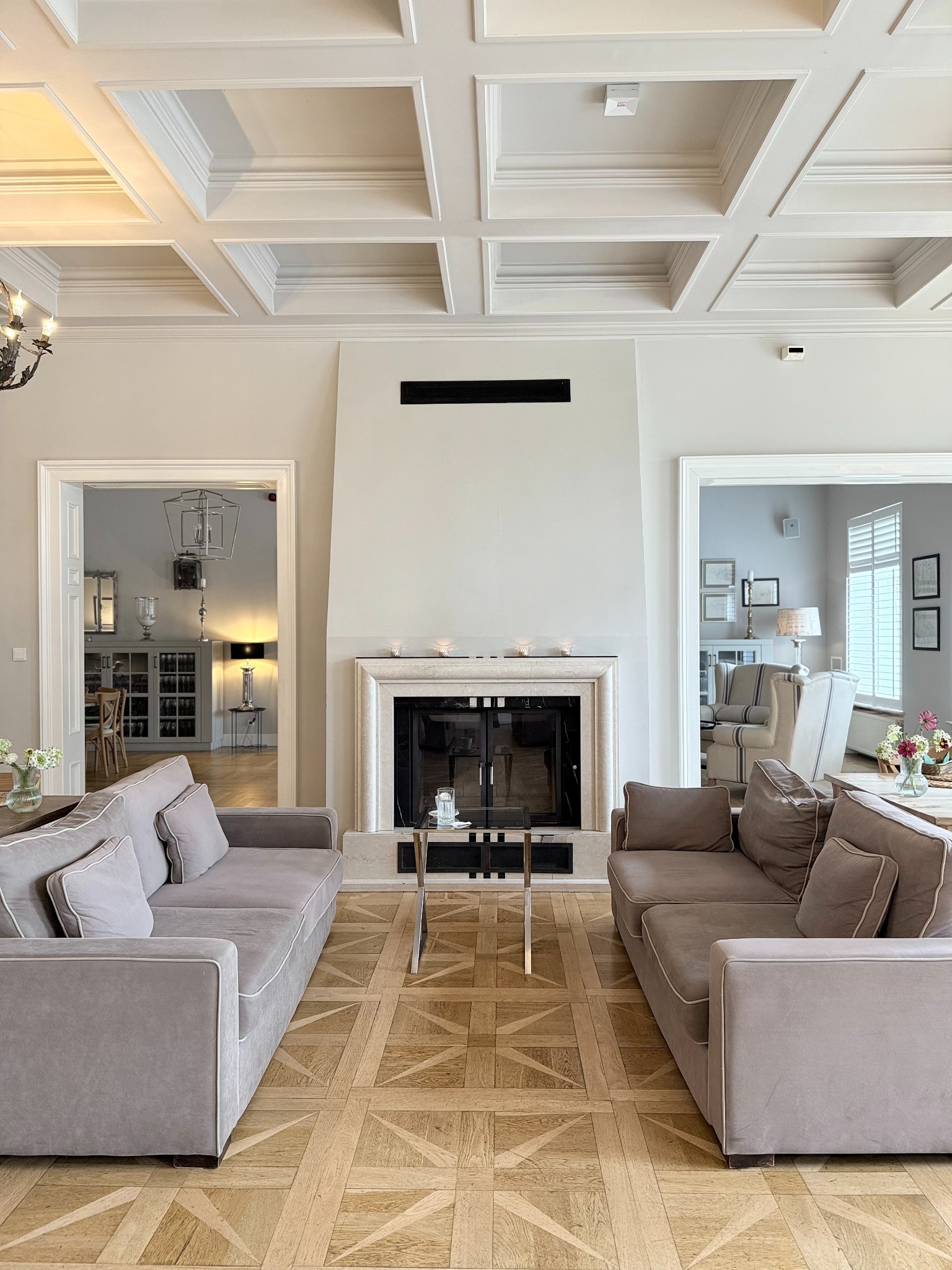 Eleganckie wnętrze salonu z symetrycznie ustawionymi sofami, kominkiem i parkietem w geometryczny wzór. Widoczne przejścia do innych pomieszczeń.