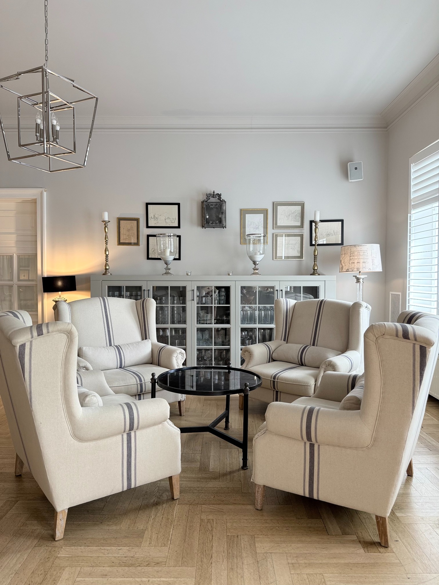 Elegancki salon z czterema fotelami w paski wokół czarnego stolika. Parkiet w jodełkę, szara komoda z dekoracjami i lampa z abażurem w tle.