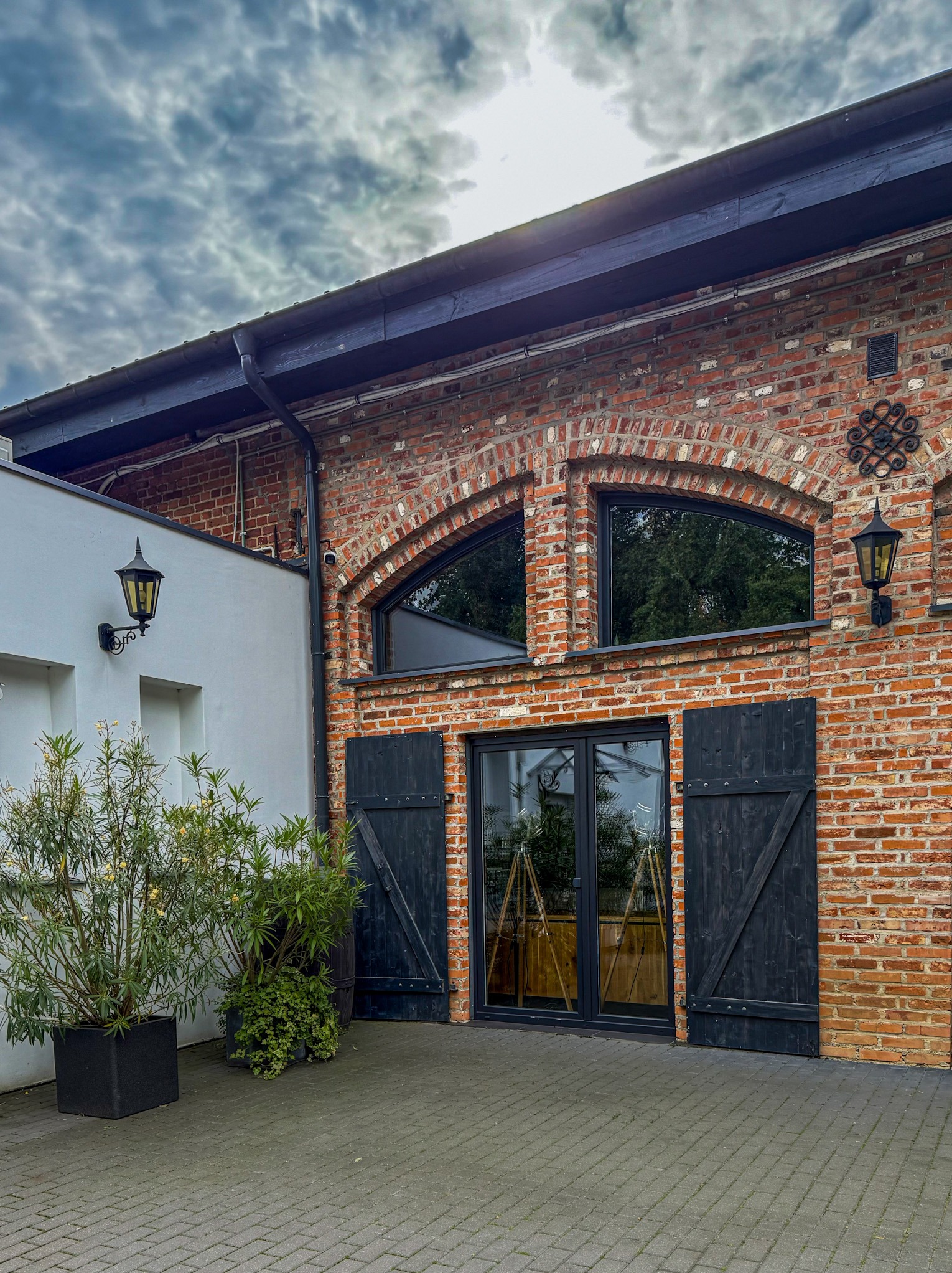 Murowana fasada budynku z cegły z czarnymi okiennicami i oknami łukowymi, oświetlona lampami. Rośliny w donicach przed wejściem.