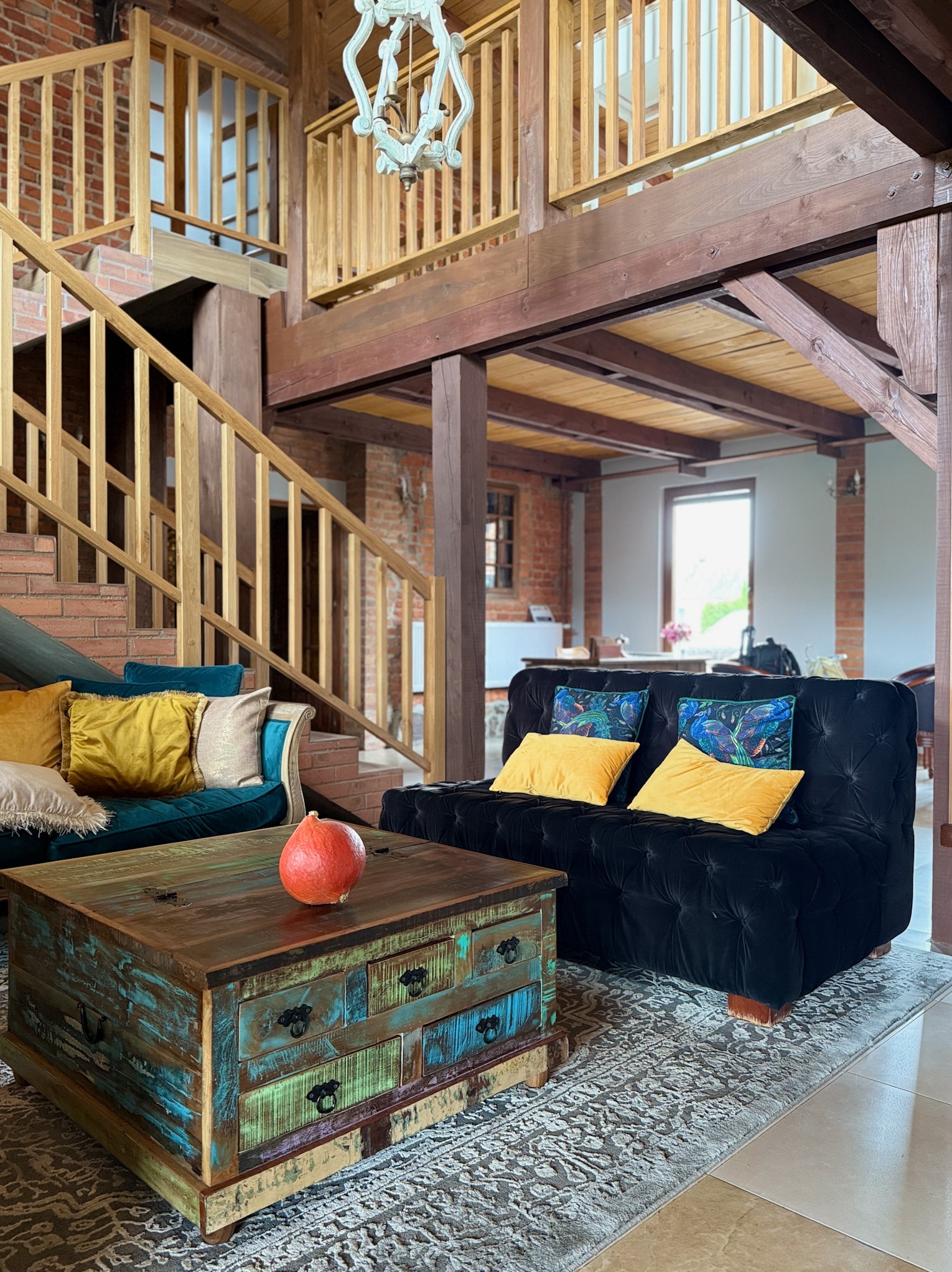Rustykalny salon z drewnianymi schodami i antresolą. Turkusowa sofa, czarna kanapa z żółtymi poduszkami, stolik kawowy z odzyskanego drewna i dekoracyjna dynia.