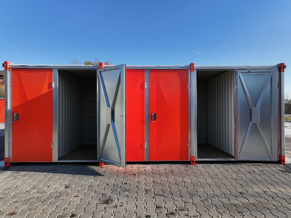 Rząd czerwono-srebrnych kontenerów magazynowych z otwartymi drzwiami na zewnątrz, widok z przodu. Kontenery ustawione na kostce brukowej pod błękitnym niebem.