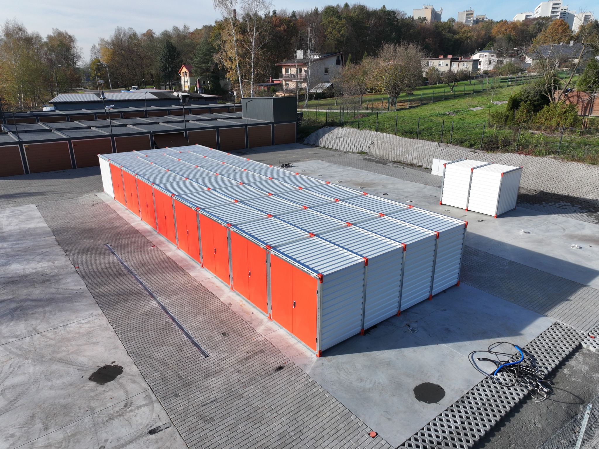 Rząd pomarańczowych i białych kontenerów magazynowych na zewnątrz, widok z góry, betonowa nawierzchnia, w tle zabudowa miejska i drzewa.