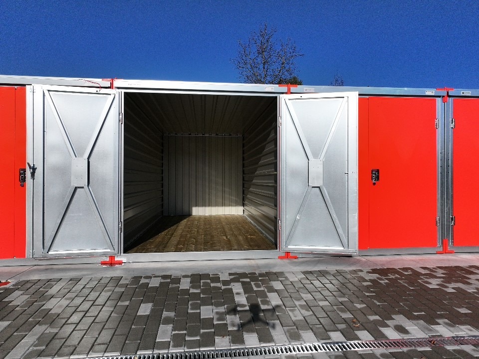 Otwarty, metalowy kontener magazynowy z czerwonymi drzwiami na zewnątrz, widok na puste wnętrze i podłogę z desek, na tle niebieskiego nieba.