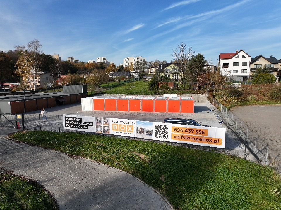 Zewnętrzny widok na magazyny samoobsługowe BOX z pomarańczowymi drzwiami, widoczny baner reklamowy z numerem telefonu i adresem strony internetowej.