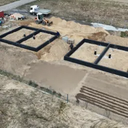 Dwie czarne, prostokątne formy fundamentowe na placu budowy, widok z góry. W tle maszyny budowlane i kontener. Częściowo ogrodzony teren.