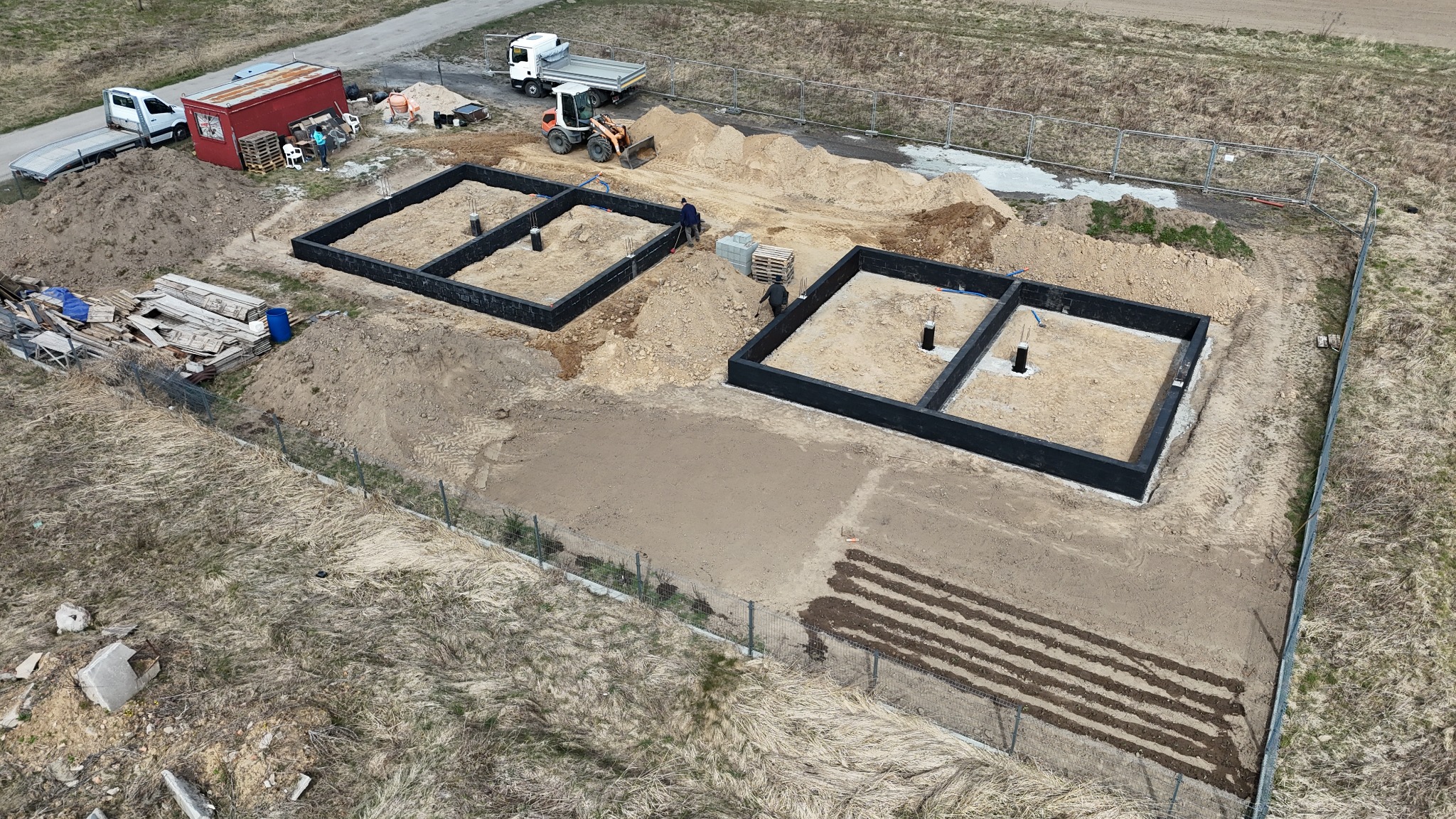Dwie czarne, prostokątne formy fundamentowe na placu budowy, widok z góry. W tle maszyny budowlane i kontener. Częściowo ogrodzony teren.