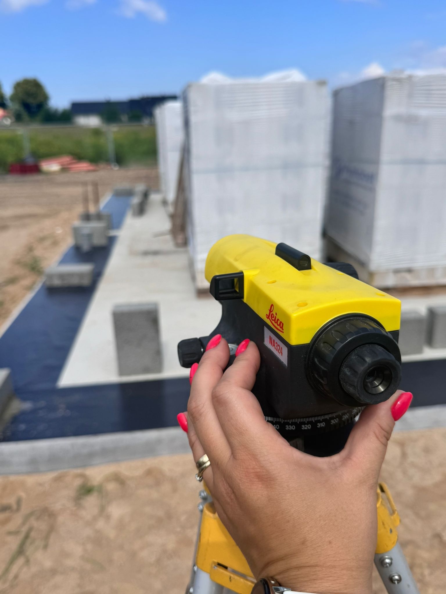 Żółty niwelator Leica NA324 w dłoni kobiety z czerwonymi paznokciami, na tle fundamentów i bloczków Ytong na budowie domu, słoneczny dzień.
