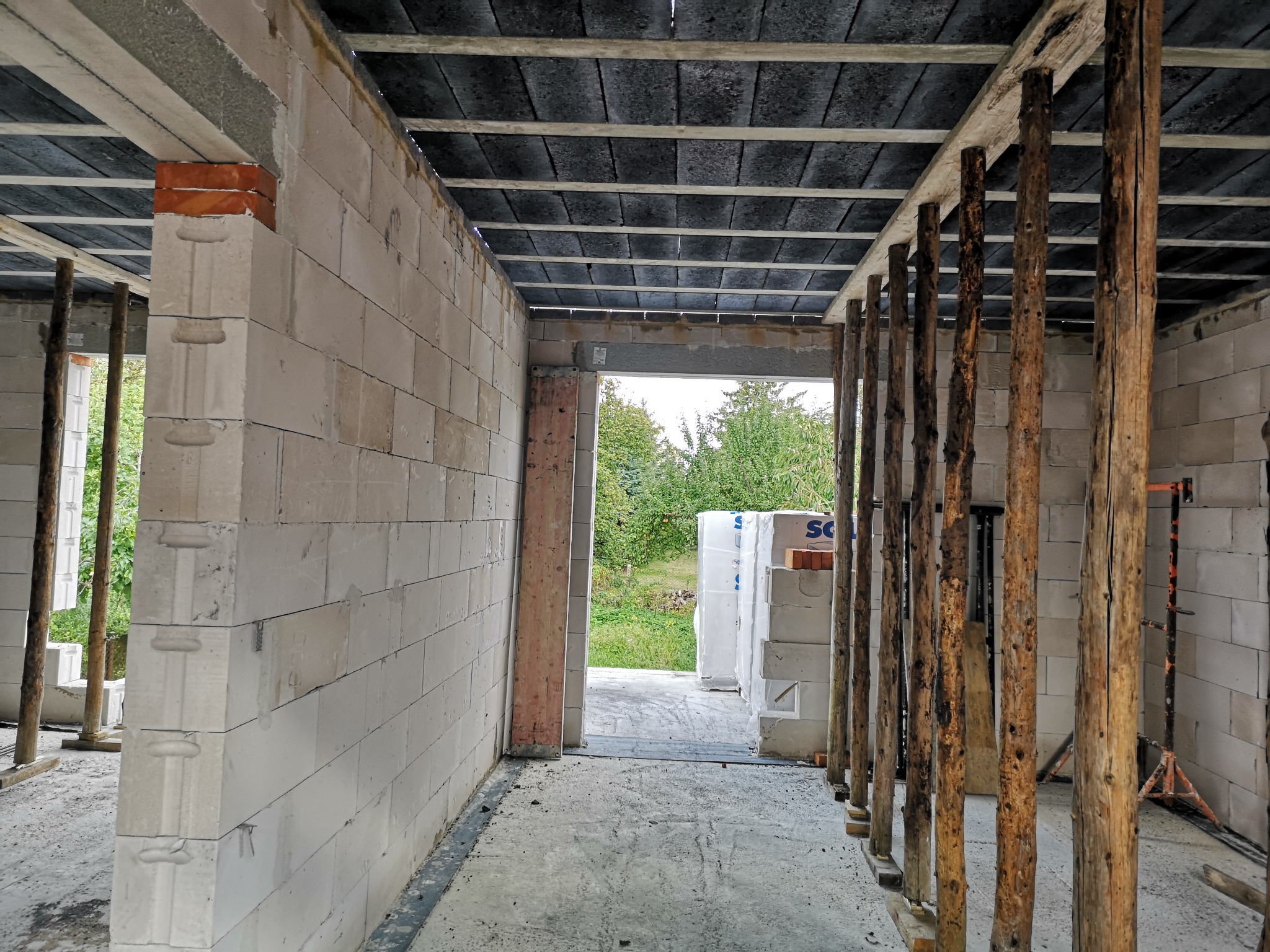 Wnętrze surowego domu w budowie z białych bloczków, drewnianymi stemplami podtrzymującymi strop, widok na zewnątrz przez otwór drzwiowy i materiały budowlane.