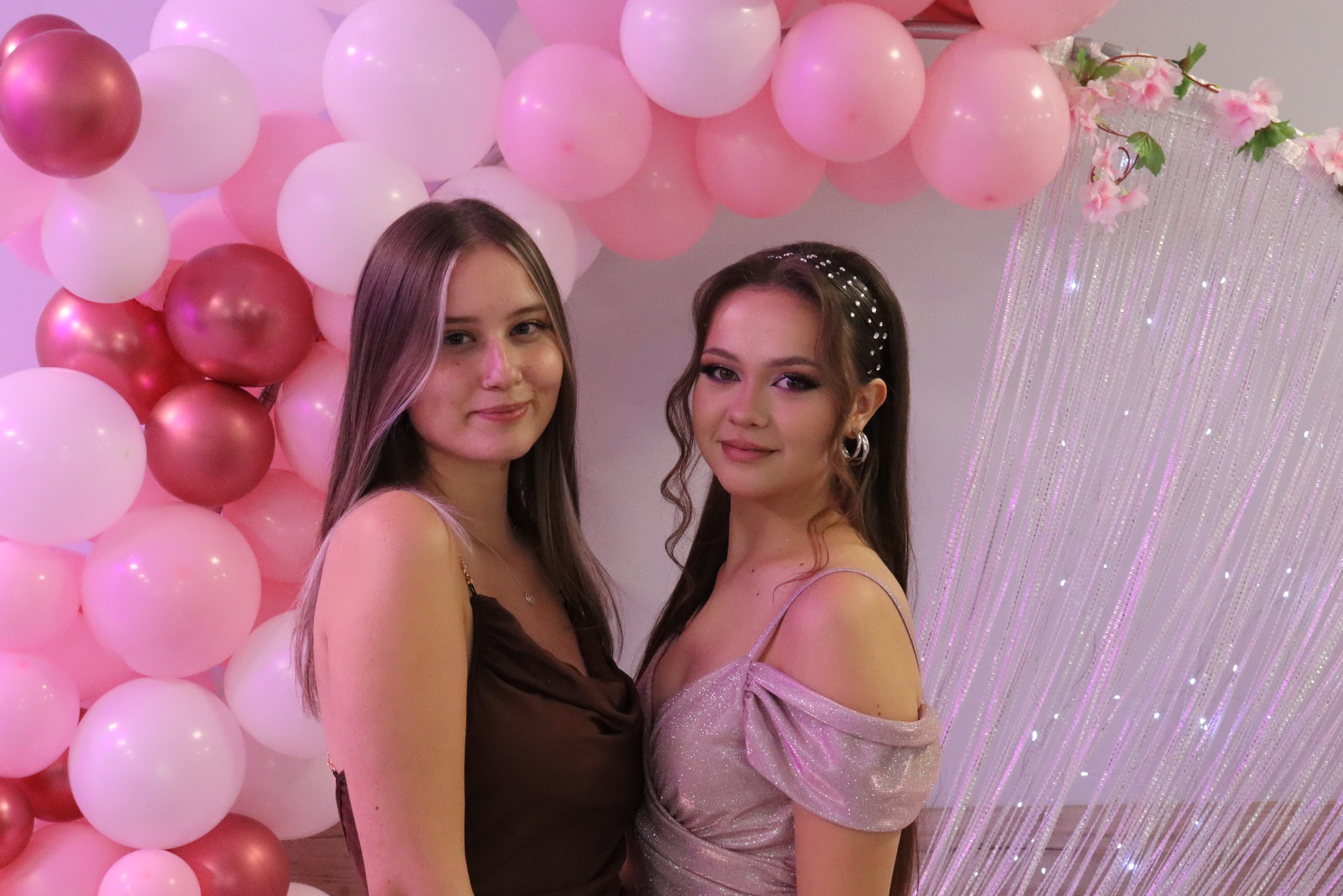 Dwie uśmiechnięte kobiety na tle dekoracji z różowych i białych balonów oraz błyszczącej ścianki, prawdopodobnie podczas sesji zdjęciowej.