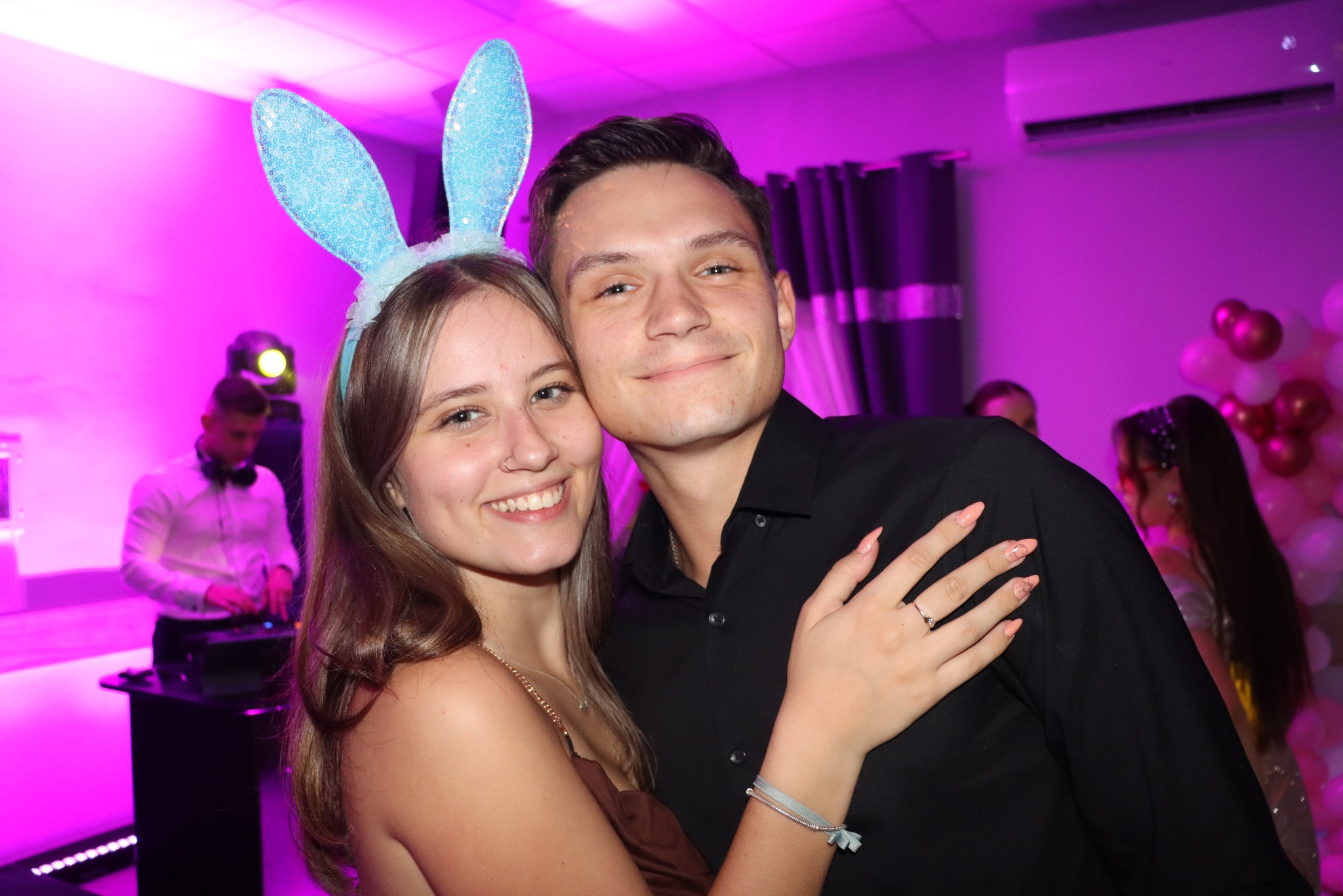 Uśmiechnięta para w Toruniu, ona z uszami królika, on w czarnej koszuli, pozuje na tle sali z dekoracjami i DJ-em, oświetlona fioletowym światłem.