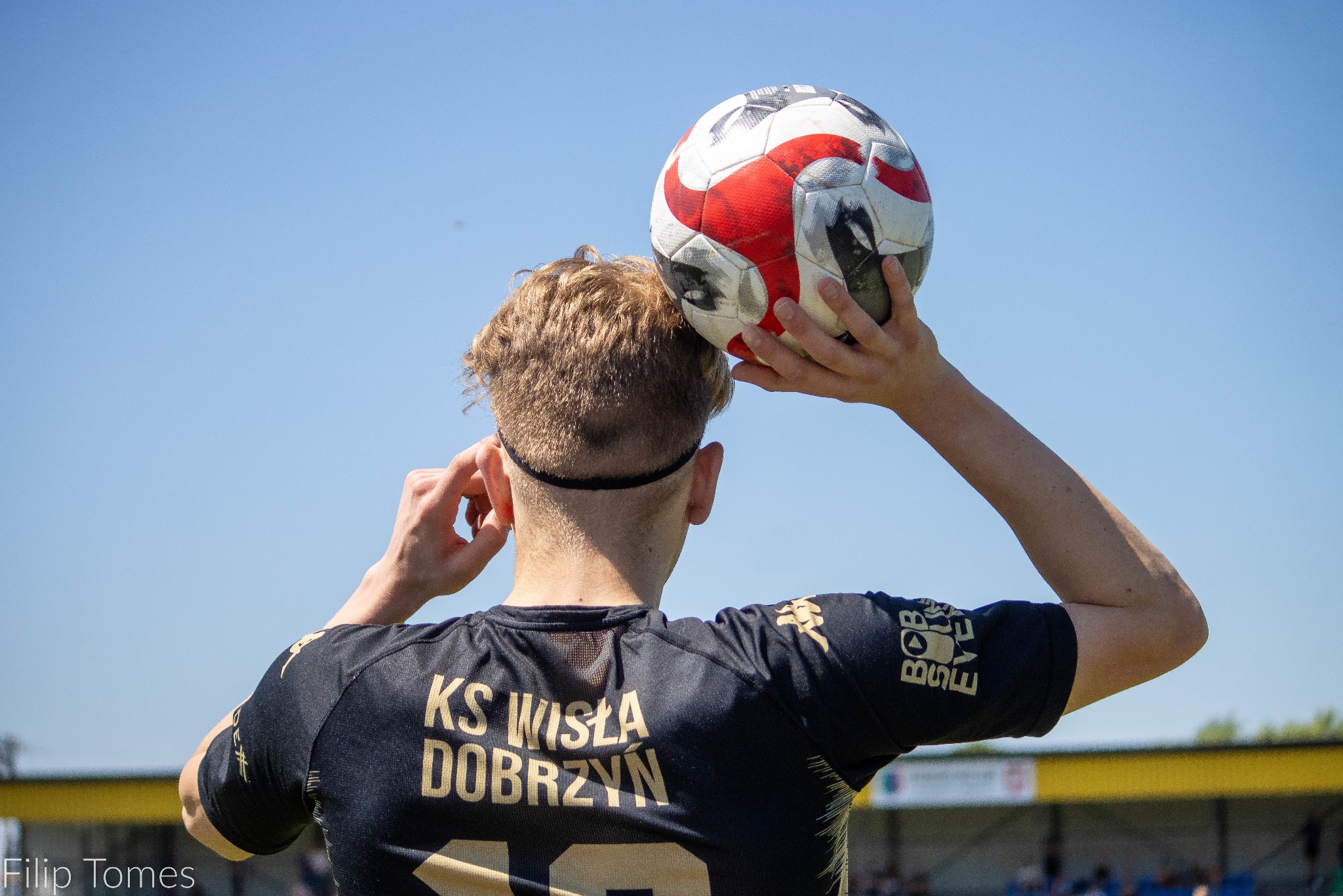 Piłkarz KS Wisła Dobrzyń z piłką na głowie, widok z tyłu, na tle błękitnego nieba i trybun. Ujęcie z boiska, w słoneczny dzień. Detal: logo sponsora na rękawie.