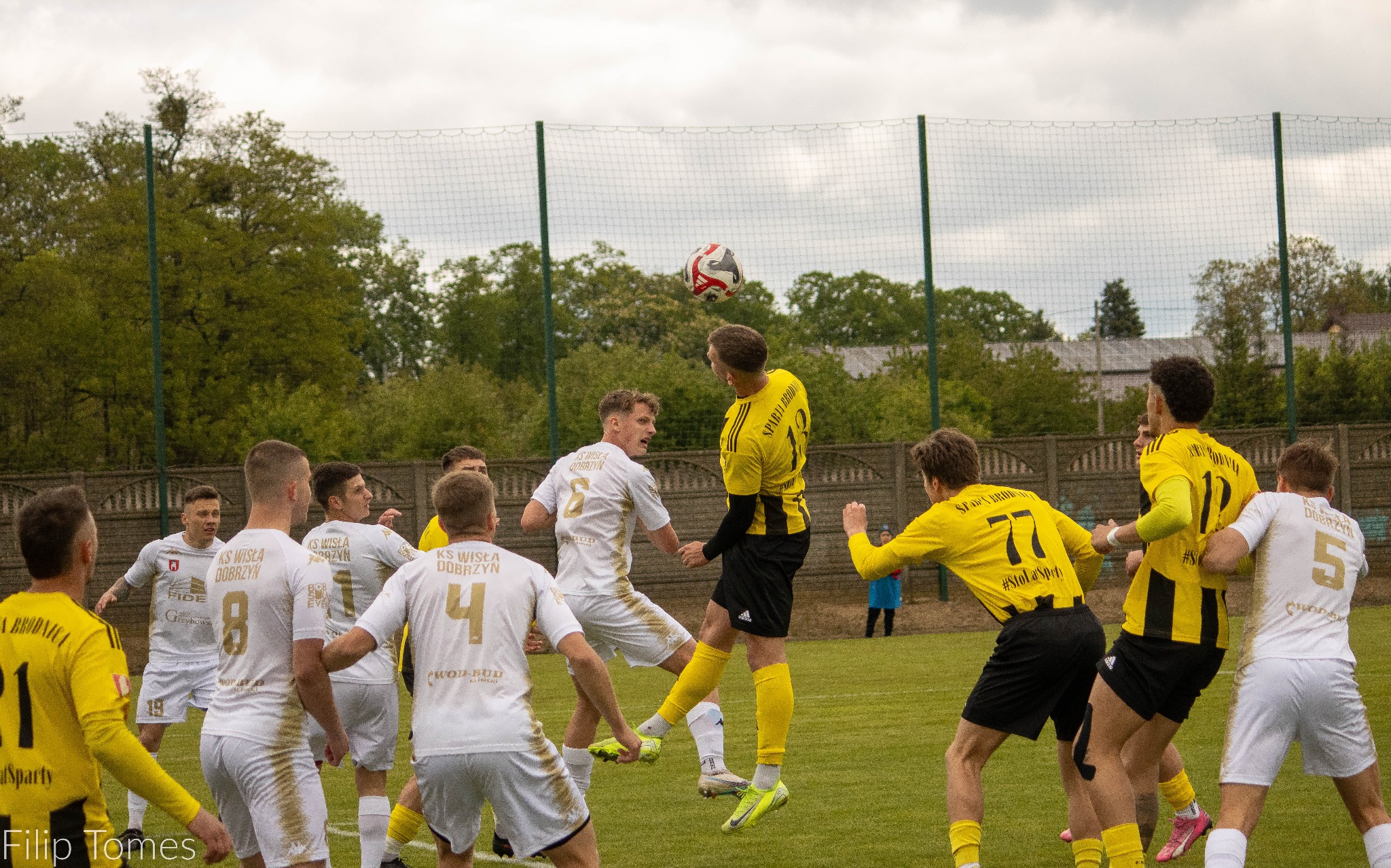 Dynamiczne ujęcie meczu piłki nożnej: zawodnicy w akcji, skok do piłki, gra zespołowa na zielonej murawie, siatka w tle i pochmurne niebo.