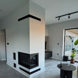 Dawid Szablewski - Nowoczesny kominek narożny z czarnymi akcentami w minimalistycznym salonie. Widoczne detale wykończenia i oświetlenie punktowe.