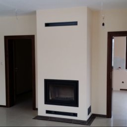 Dawid Szablewski - Nowoczesny kominek z czarnym wkładem, w jasnym salonie z widocznymi schodami i otwartymi drzwiami. Minimalistyczny design, neutralna kolorystyka ścian i podłóg.