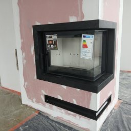 Dawid Szablewski - Nowoczesny kominek z czarną obudową w trakcie montażu, widoczne naklejki informacyjne na szybie, surowy stan wykończenia ścian i podłogi, instalacja CO w tle.